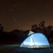 A tent, lit from within in a dusky desert scene. Joshua trees are visible against the backdrop of the starry night sky
